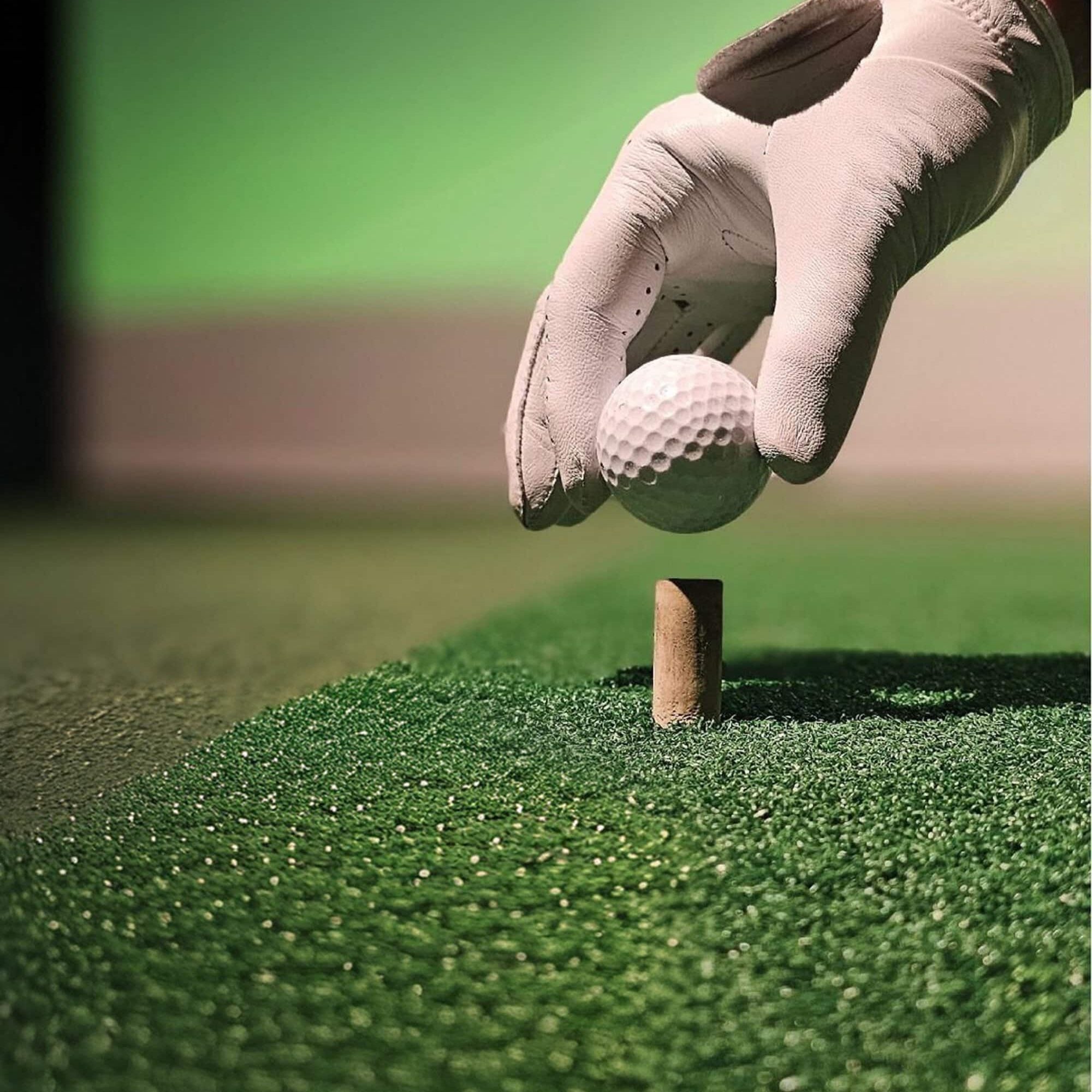 A gloved hand places a golf ball on a wooden tee set in artificial turf with a blurred green background.